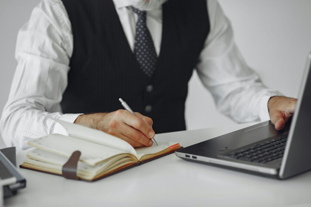 Business professional writing notes while working on a laptop at an office desk.