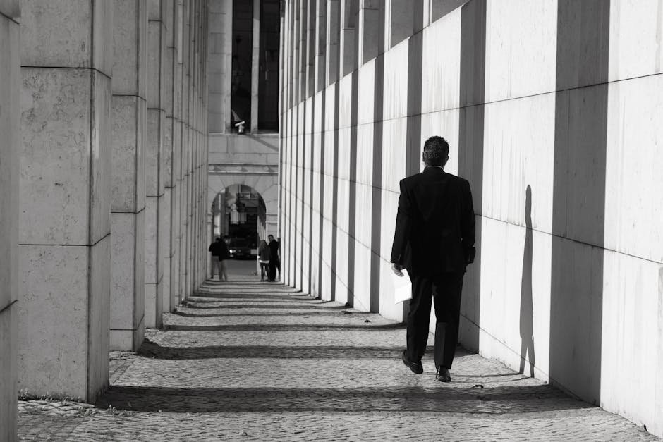 A man walks along a sunlit, shadowed sidewalk in Lisbon, Portugal.