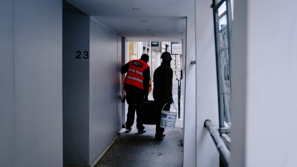 Two construction workers carrying materials in a city hallway.
