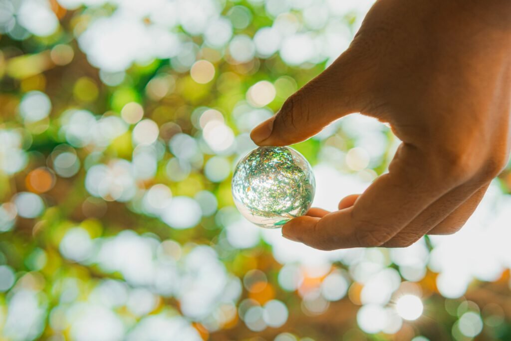 Close-up of a hand holding a glass sphere reflecting green leaves and sunlight.