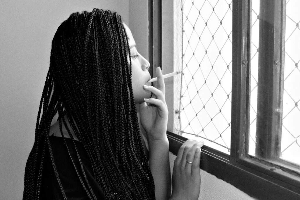 Black and white of side view of young female with braids smoking while standing near window